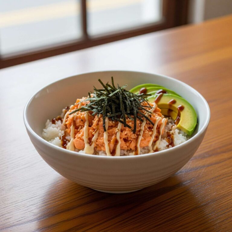 Emily Mariko's Salmon Rice Bowl on a wooden table, ready for cooking. The bowl contains mashed salmon and rice, garnished with avocado, nori sheets, and sauces.