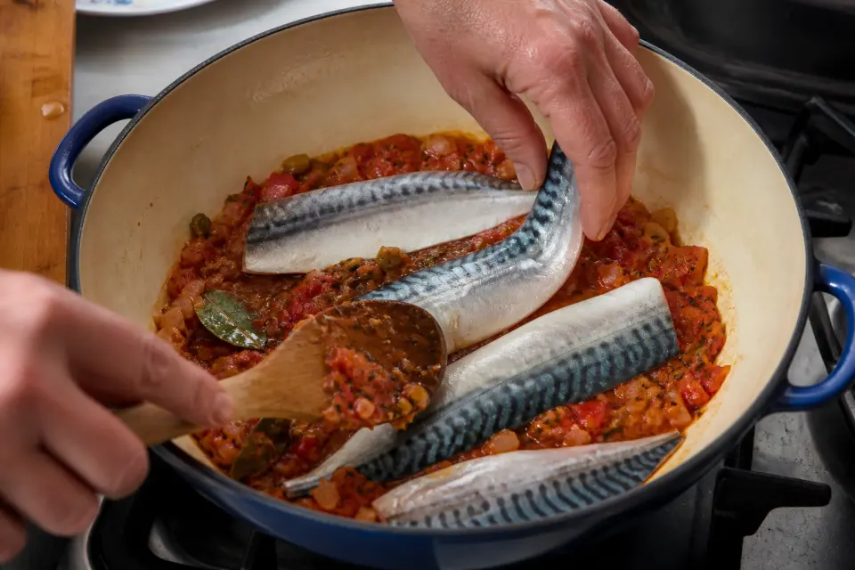 Mackerel fillets being layered with tomato and pepper sofrito in a wide pot
